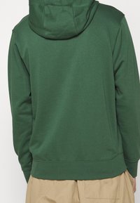 Grön hoodie med en stor, justerbar huva och ribbade fållar. Tyget verkar mjukt och strukturerat, kompletterat med en avslappnad passform.