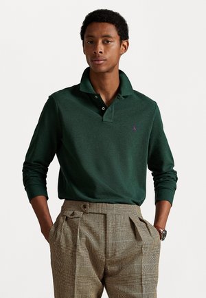 Dunkelgrünes Hemd mit langen Ärmeln und Kragen sowie lila Logo, kombiniert mit strukturierten karierter Hose mit Falten und einem Gürtel.