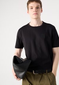 Svart t-skjorte med korte ermer, vertikale sømmer og et firkantet logomerke. Modellen holder en svart, strukturert veske. Olivengrønne bukser er synlige.