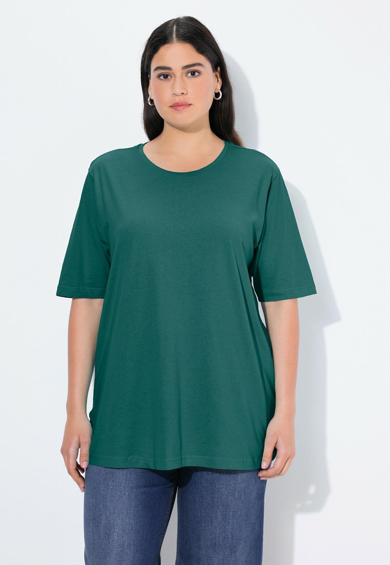 Frau mit langen dunklen Haaren, die silberne Creolenohrringe, ein dunkelgrünes Kurzarmshirt und blaue Jeans trägt, steht vor einem weißen Hintergrund.