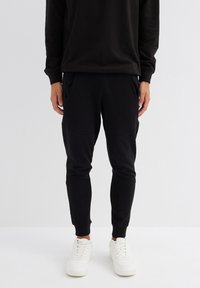 Pantalones de chándal negros con piernas ajustadas, cinturilla elástica y dos bolsillos laterales. Combinados con zapatillas blancas. Textura de tela suave.