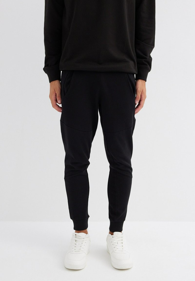 Pantalones de chándal negros con piernas ajustadas, cinturilla elástica y dos bolsillos laterales. Combinados con zapatillas blancas. Textura de tela suave.