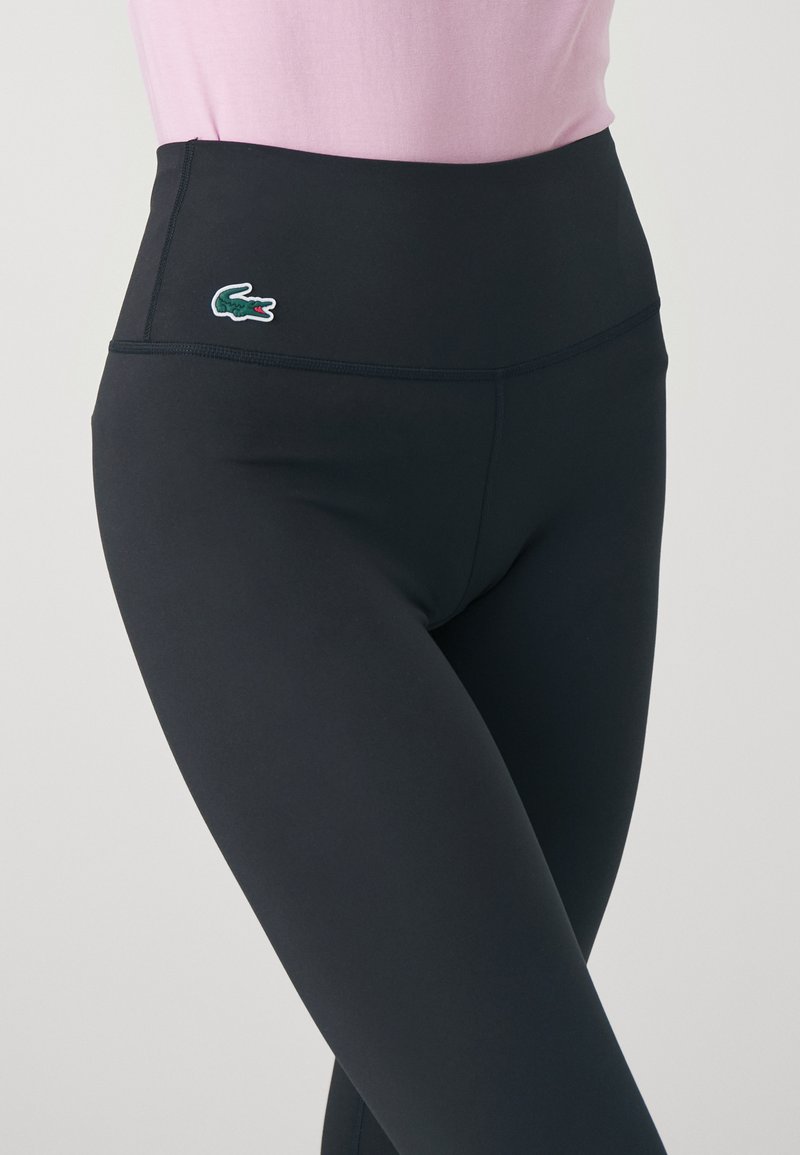 Leggings neri a vita alta con una texture liscia, caratterizzati da un logo Lacoste verde e bianco sulla cintura.