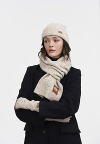 Kvinde iført en sort frakke, beige strikket hat med striber, matchende tørklæde omkring halsen og fingerløse handsker, der ser til siden.