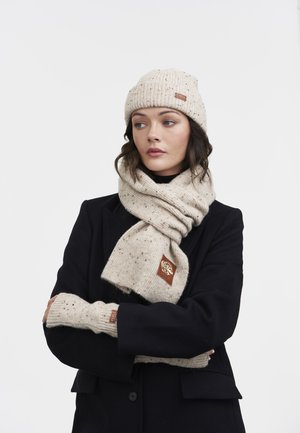 Femme portant un manteau noir, un bonnet en tricot beige à pois, une écharpe assortie enroulée autour du cou et des gants sans doigts, regardant sur le côté.