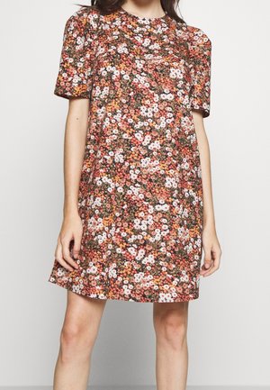 Kobieta ubrana w sukienkę do kolan z krótkim rękawem, o gęstym kwiatowym wzorze w odcieniach różu, pomarańczu, bieli i zieleni na brązowym tle.