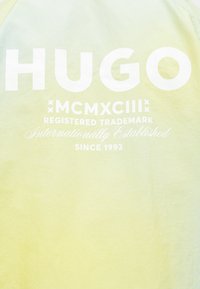 Κίτρινο ύφασμα με έντονο λευκό κείμενο "HUGO", ρωμαϊκούς αριθμούς MCMXCIII και τη φράση "Καταχωρημένο εμπορικό σήμα διεθνώς καθιερωμένο από το 1993."