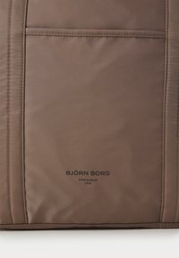 Smeđa platnena torba s prednjim džepom, glatke teksture, prošivenim detaljima i utisnutim logom s natpisom "BJÖRN BORG STOCKHOLM 1956".