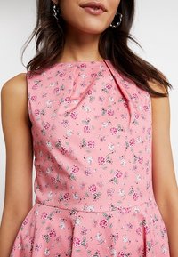 Žena v bezramínkových růžových šatech s malými růžovými a bílými květinovými vzory a sklady v oblasti výstřihu a pasu.