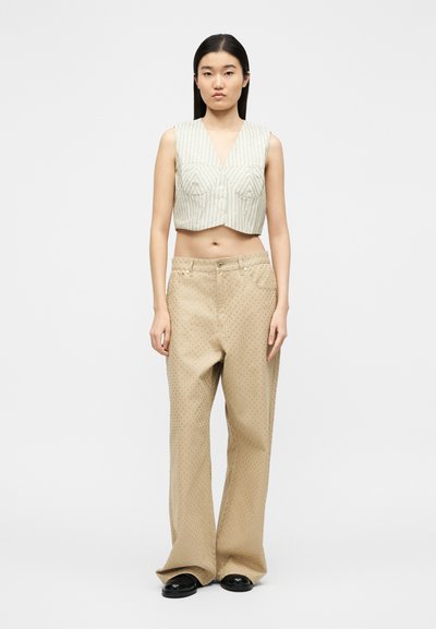 Γυναίκα που φορά αμάνικο κροπ μπεζ ριγέ γιλέκο και μπεζ παντελόνι με φαρδιά μπατζάκια και μικρές οπές, στέκεται μπροστά σε απλό φόντο.