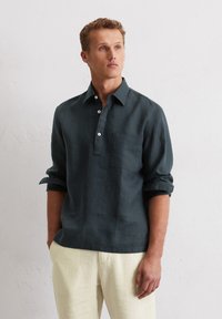 Chemise en lin vert foncé avec une patte de boutonnage, manches longues retroussées et une poche poitrine, associée à un pantalon beige clair.