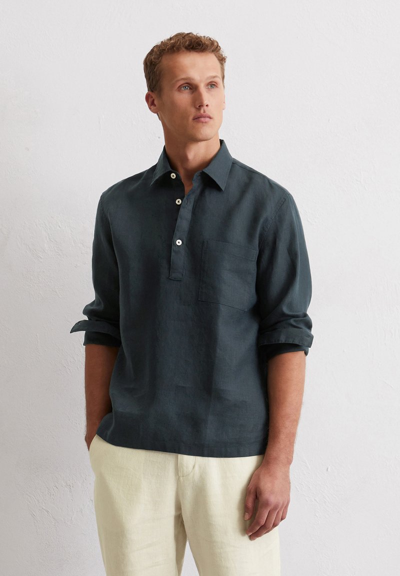 Chemise en lin vert foncé avec une patte de boutonnage, manches longues retroussées et une poche poitrine, associée à un pantalon beige clair.