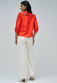 Blusa roja de manga corta con detalles de encaje en los hombros, combinada con pantalones anchos blancos y sandalias de tacón beige. Textura suave.