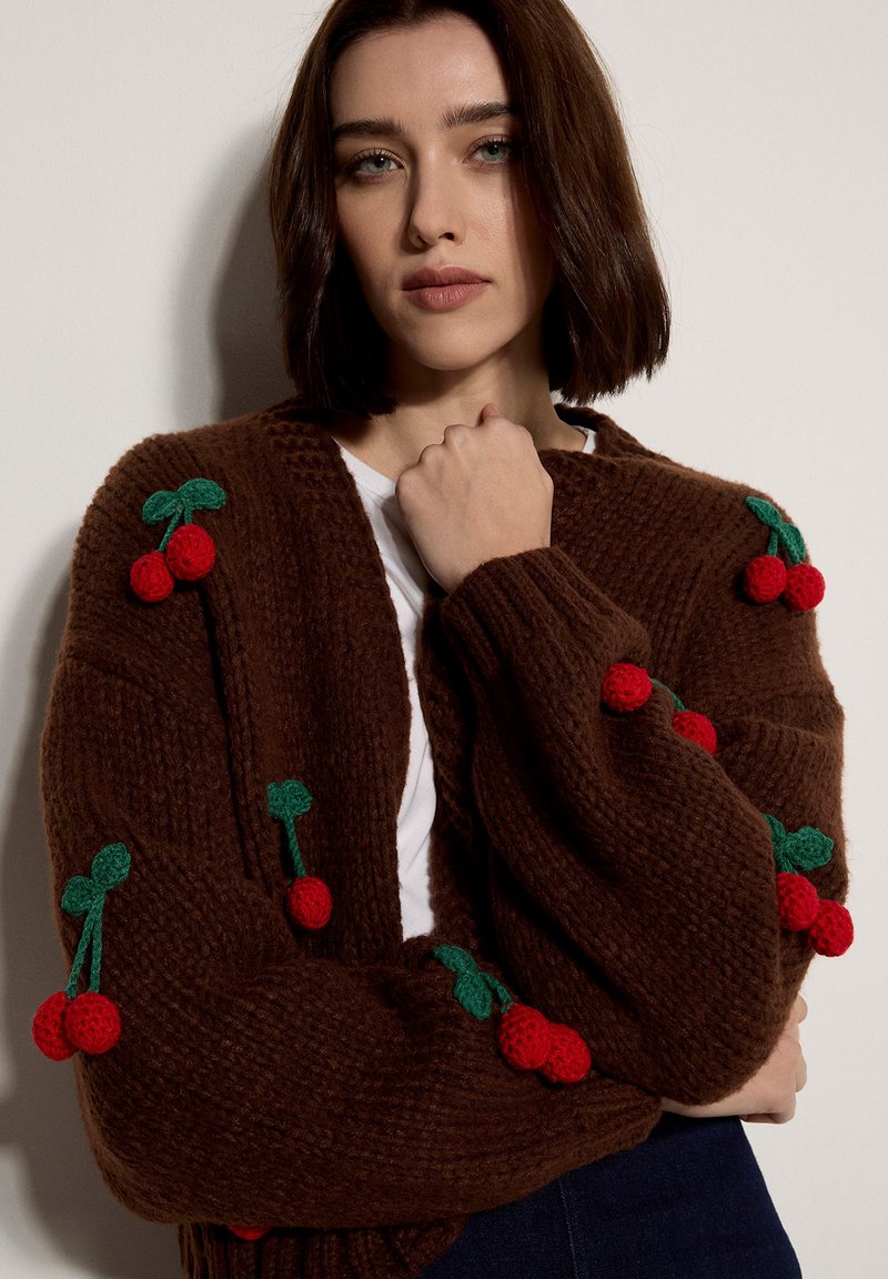 Jeune femme aux cheveux bruns mi-longs portant un cardigan en tricot marron orné de cerises rouges et de feuilles vertes.