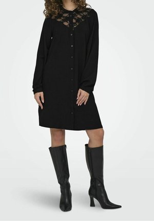 Woman wearing a black button-up dress with lace details at the top and knee-high black heeled boots, standing with hands relaxed.