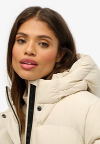 Beige hættejakke med dunfyld, lukkes med lynlås og knapper, lavet af struktureret stof med sorte detaljer langs lynlåsen.