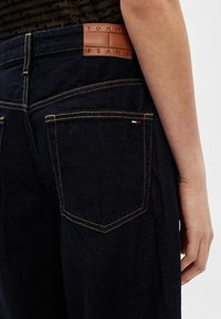 Κοντινό πλάνο σκούρων μπλε τζιν με ραφές σε καφέ απόχρωση, καφέ δερμάτινο πατς "Tommy Jeans" και ένα μικρό κόκκινο, λευκό και μπλε λογότυπο κοντά στην πίσω τσέπη.