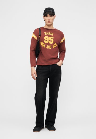 Jeune homme portant une chemise à manches longues bordeaux avec un texte jaune, pantalon noir, casquette noire, sac à bandoulière noir, debout avec une main dans la poche.