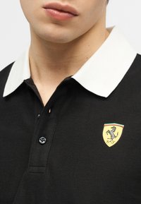 Gros plan d'une personne portant un polo noir avec un col blanc et le logo Ferrari, représentant un cheval cabré et les initiales "S F".
