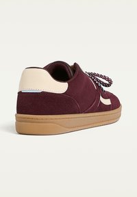 Bordeaux ruskindsneaker med en cremefarvet hælaccents, tekstureret overdel og mønstrede snørebånd, sat på en gummisål med et lavprofil design.