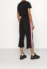 Czarne spodnie o kroju cropped z wysokim stanem, posiadające wielokolorowe paski po bokach w odcieniach różu, fioletu i kremu. Połączone z czarnymi trampkami.