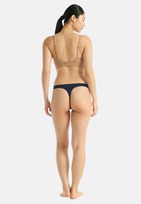 Eine Frau steht barfuß mit dem Rücken zur Kamera, trägt einen beigen BH und marineblaue Strings vor einem schlichten hellen Hintergrund.