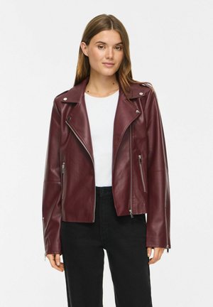 Jeune femme portant une veste en cuir bordeaux par-dessus une chemise blanche et un pantalon noir, debout devant un fond blanc uni.