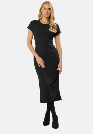 Vrouw met lang blond haar, gekleed in een nauwsluitende zwarte midi-jurk met korte mouwen, gecombineerd met zwarte panty's en zwarte hakken, die zelfverzekerd staat.