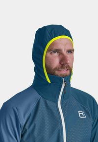 Blue softshell jacket with high collar and attached hood. Features bright yellow accents along the hood. Zippered front and textured pattern on fabric.