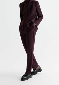 Burgundský oblek na míru s vypasovanou bundou, spárovaný s koženými úzkými kalhotami s postranními rozparky. Nošený s černými robustními mokasínami.