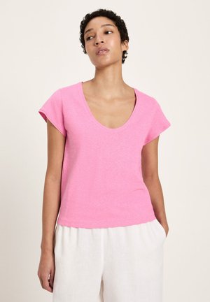 Frau trägt ein rosa T-Shirt mit rundem Ausschnitt und kurzen Ärmeln sowie weiße Hosen, steht mit einer Hand in der Tasche vor einem schlichten Hintergrund.