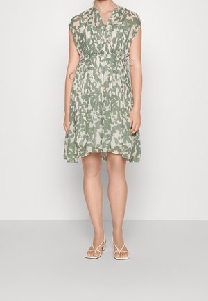Vrouw draagt een knielange jurk met groene en witte camouflageprint, cap sleeves en witte sandalen met hak en bandjes.