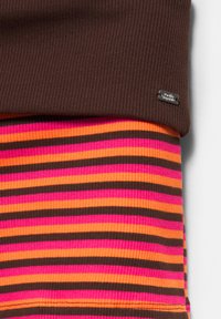 Gros plan des couches de tissu côtelé : maille marron au-dessus de rayures horizontales roses, orange et marron, avec une petite étiquette métallique rectangulaire de la marque.