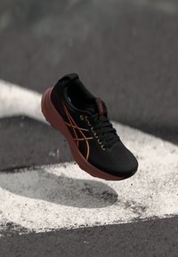 Chaussure de sport noire avec une tige en mesh, accents orange et jaune, un col rembourré et une semelle amortissante, présentée au-dessus d'une ligne blanche.