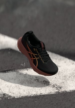 Chaussure de sport noire avec une tige en mesh, accents orange et jaune, un col rembourré et une semelle amortissante, présentée au-dessus d'une ligne blanche.