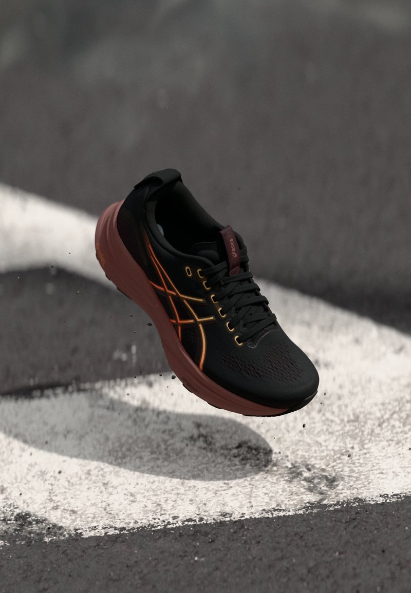 Chaussure de sport noire avec une tige en mesh, accents orange et jaune, un col rembourré et une semelle amortissante, présentée au-dessus d'une ligne blanche.