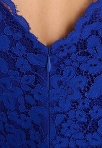 Detailní záběr na královsky modrou krajkovou látku s květinovými vzory a středním zipem na holé kůži.
