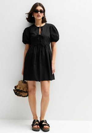Black short dress with puff sleeves, tie detail at neckline, and gathered waist. Paired with black platform sandals and a woven bag.