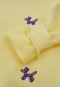 Felpa gialla con una texture morbida, caratterizzata da un design di animale a palloncino ricamato in blu e arancione sul davanti e polsini lavorati a maglia.