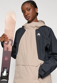 Bežna in črna vodoodporna jakna z zadrgo spredaj, kapuco in logotipom. Model drži smučko s rožnato-rjavo zasnovo in vezmi.