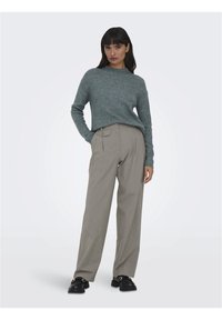 Pull en maille côtelée gris associé à un pantalon beige plissé, avec un détail de bouton. Le modèle porte des chaussures noires à semelles épaisses. Fond simple.