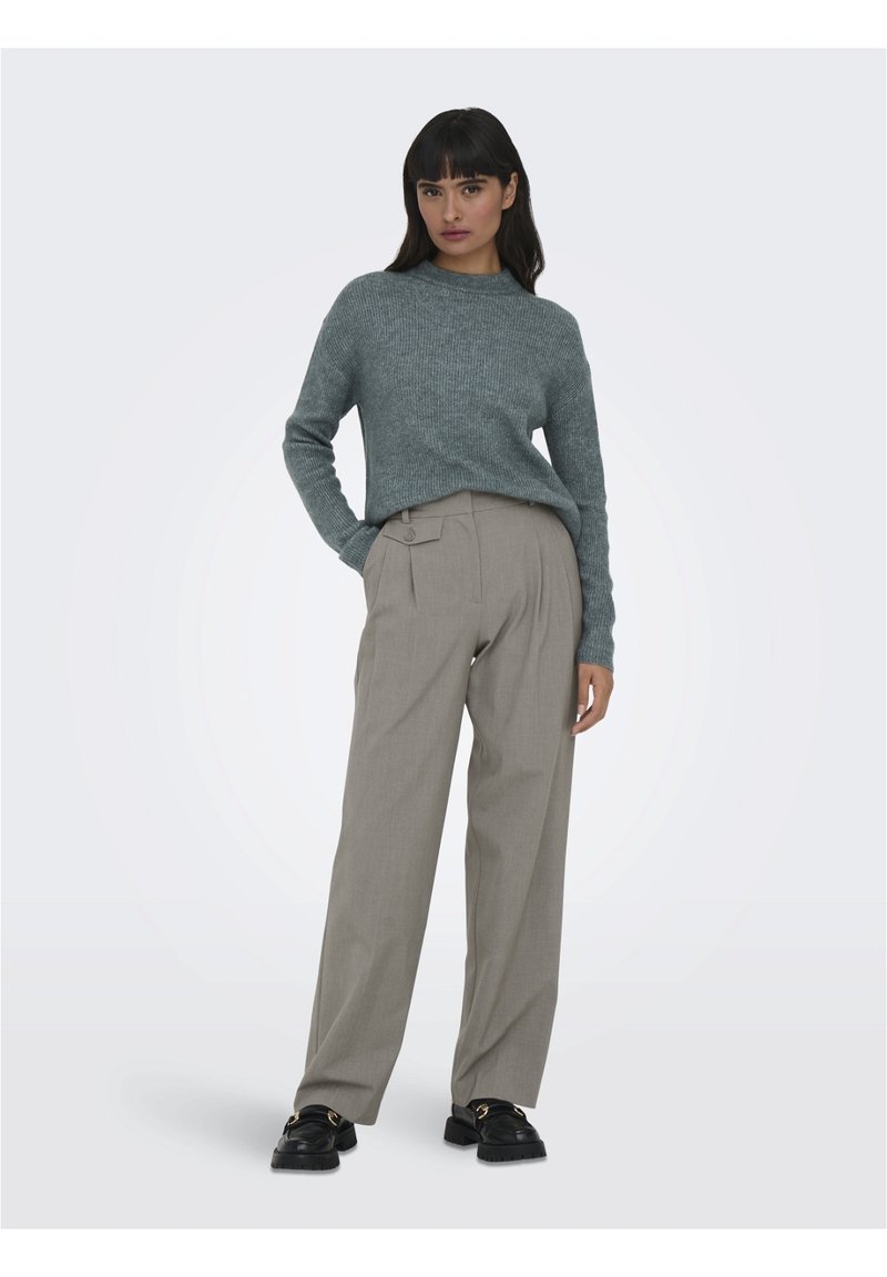 Maglione grigio a coste abbinato a pantaloni beige plissettati, con un dettaglio di bottoni. Il modello indossa scarpe nere chunky. Sfondo semplice.