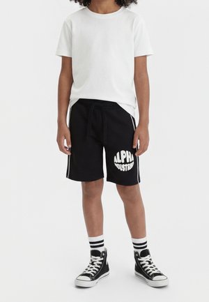 Dziecko ubrane w gładką białą koszulkę z krótkim rękawem, czarne szorty z logo „Alpha Industries”, paskowane skarpetki i czarne wysokie trampki, stoi.
