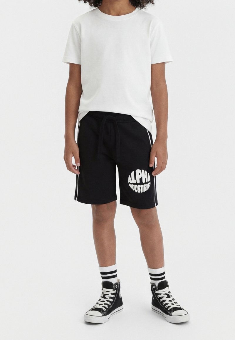 Enfant portant un t-shirt blanc uni à manches courtes, un short noir avec le logo « Alpha Industries », des chaussettes rayées et des baskets montantes noires, debout.