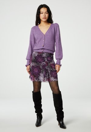 Paarse gebreide cardigan met een V-hals en knopen, gecombineerd met een paarse rok met bloemenpatroon. Model draagt kniehoge zwarte suède laarzen.