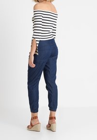 Žena v černobílém pruhovaném topu s nastavitelnými ramínky, ve vysokých modrých džínech a hnědých klínových sandálech, stojí s částečně otočeným zády.