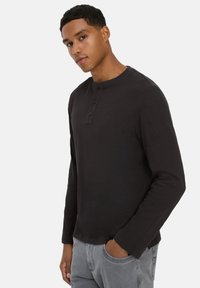 Sort langærmet henley-shirt med tre knapper ved kraven, ribbet tekstur, rund kant og et subtilt logo på venstre ærme.