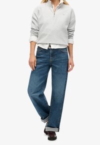 Sudadera gris con cremallera sobre una camisa blanca, combinada con vaqueros azules de pierna ancha y zapatos oscuros, con puños remangados y un ajuste holgado.