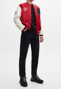 Veste universitaire rouge et crème avec détails brodés, chemise noire, jean foncé et baskets noires avec accents gris.