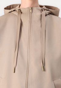 Casaco bege com capuz, zíper frontal, cordões e bolsos laterais. Feito de um tecido liso e leve com detalhes de costura subtis.